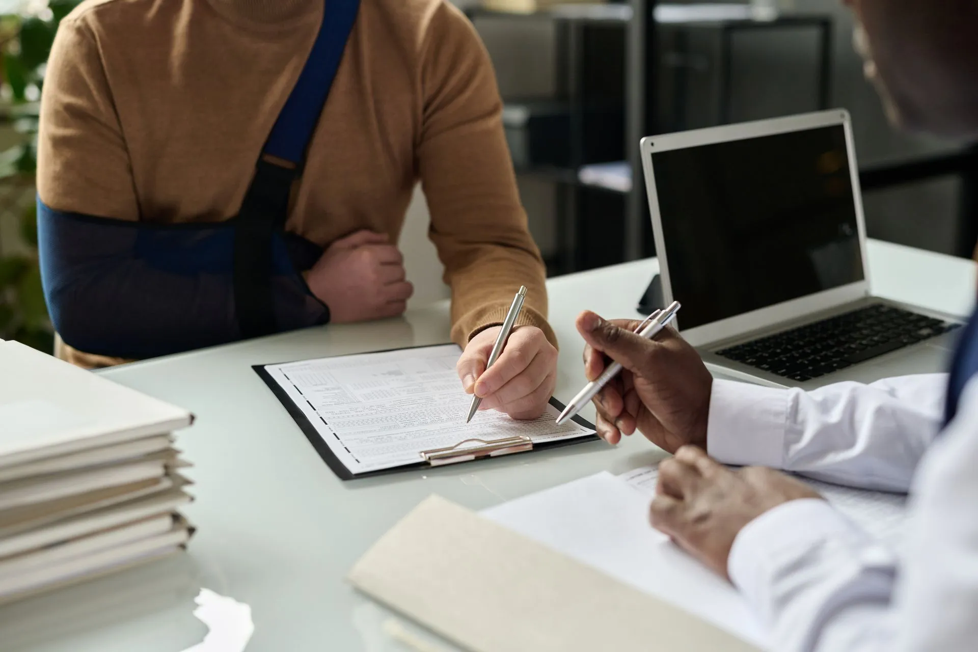 Person with arm in sling filling out a form while consulting a professional across a desk with a laptop.