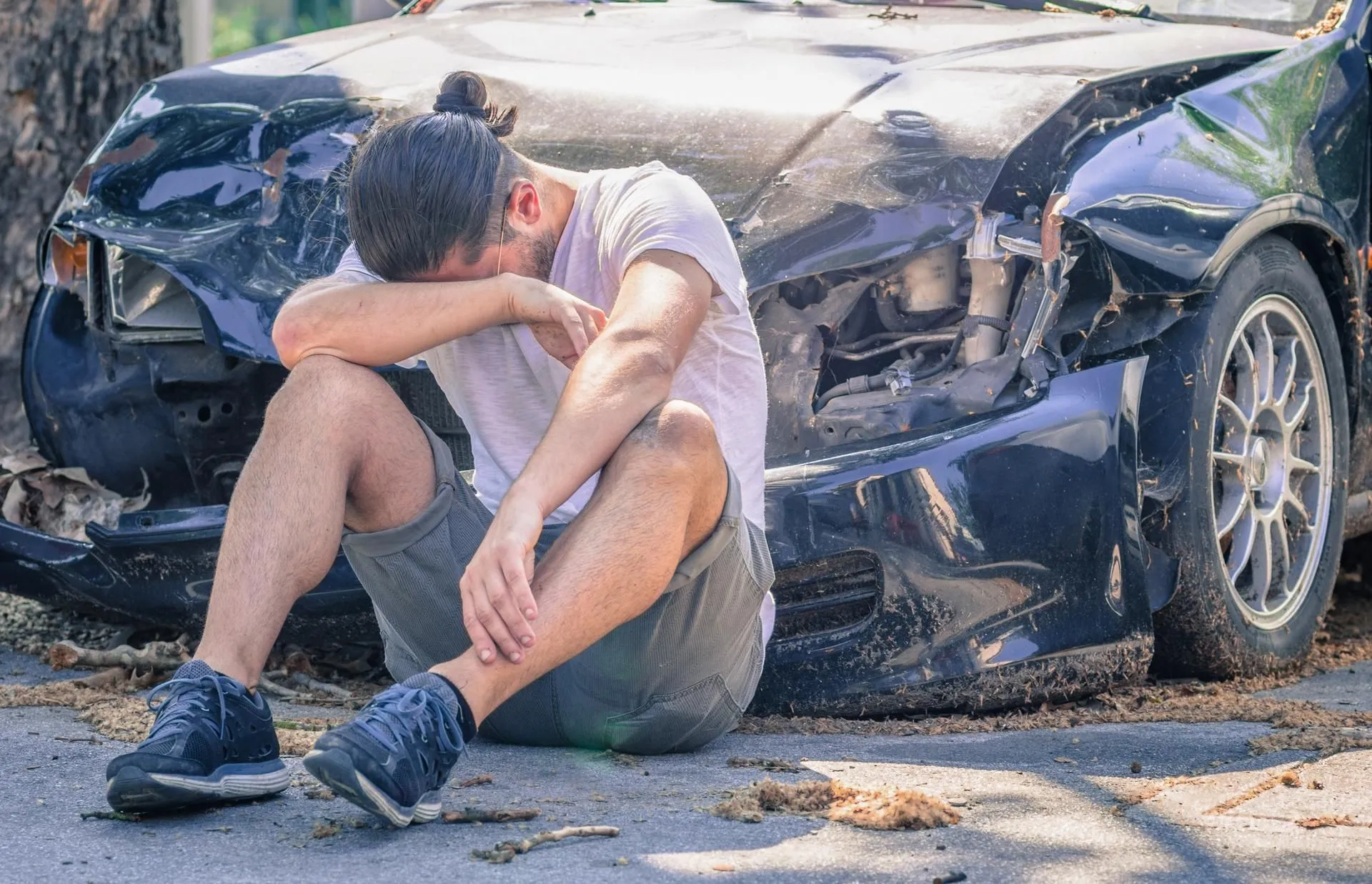 A distressed man sitting in front of a heavily damaged car.