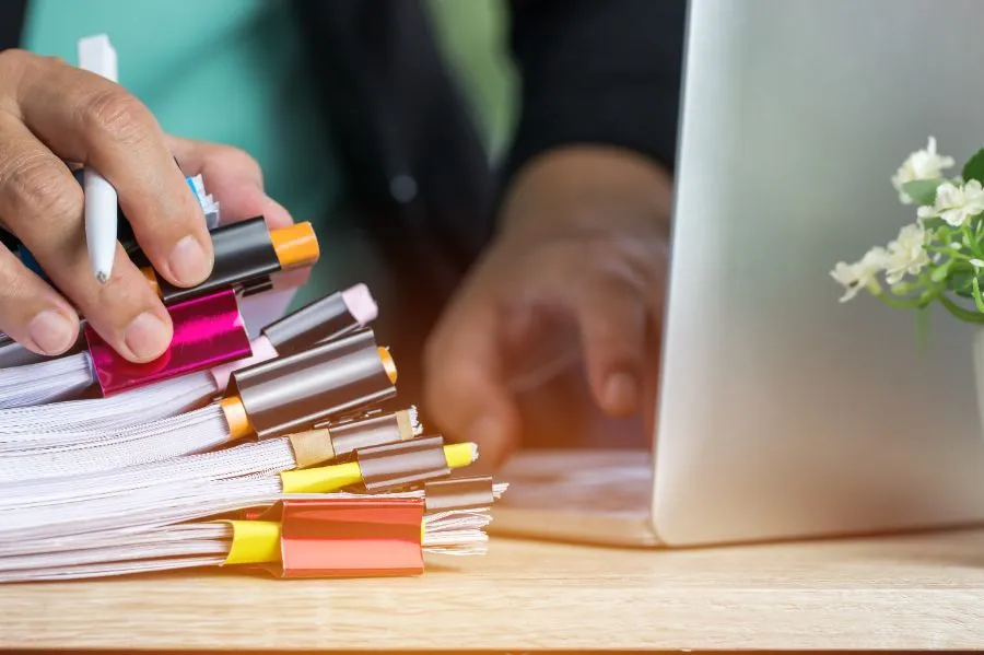 Person's hands holding a pen on a thick stack of clipped papers next to a laptop.