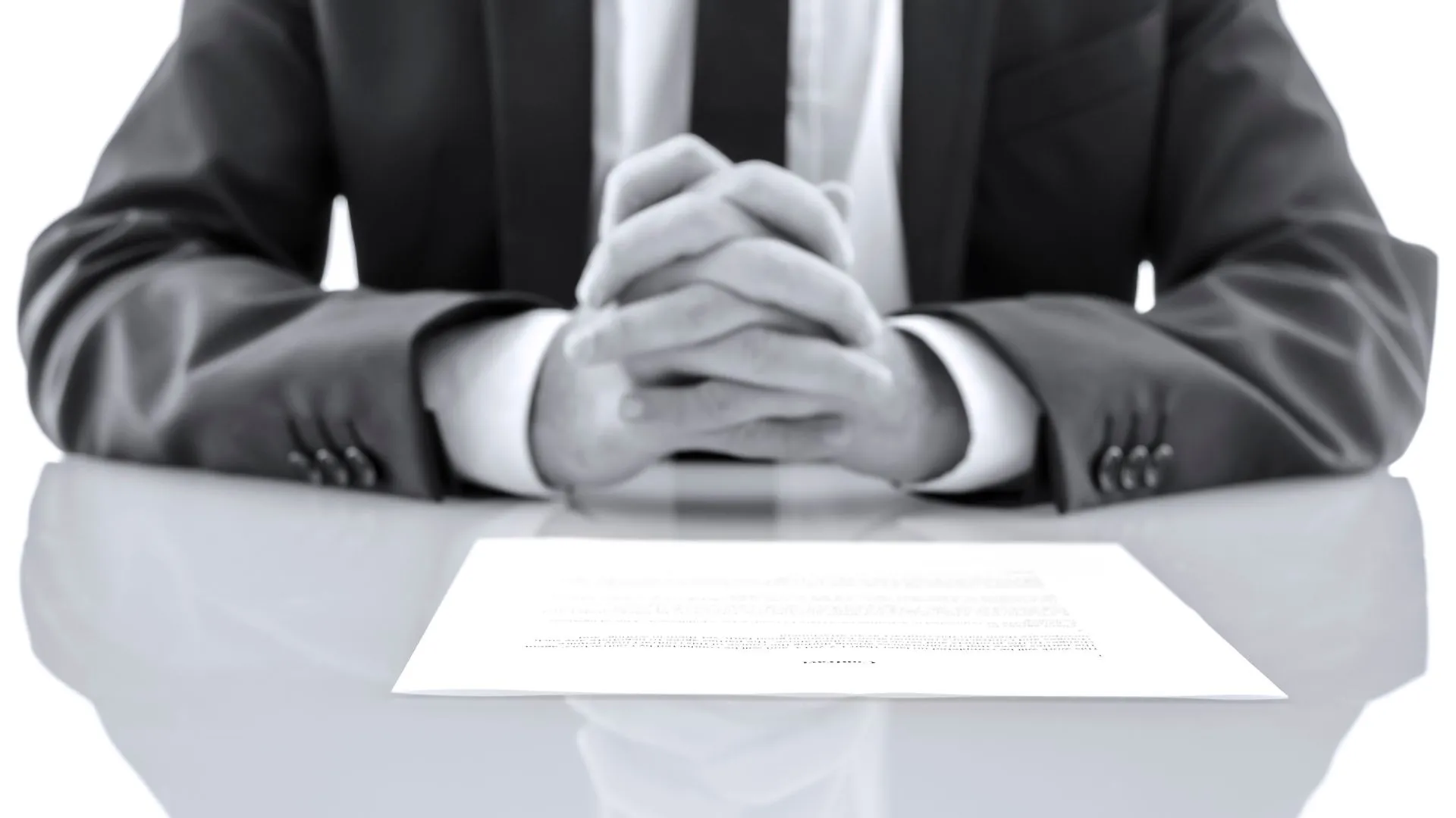 Person in a suit with clasped hands sitting at a desk behind a document.