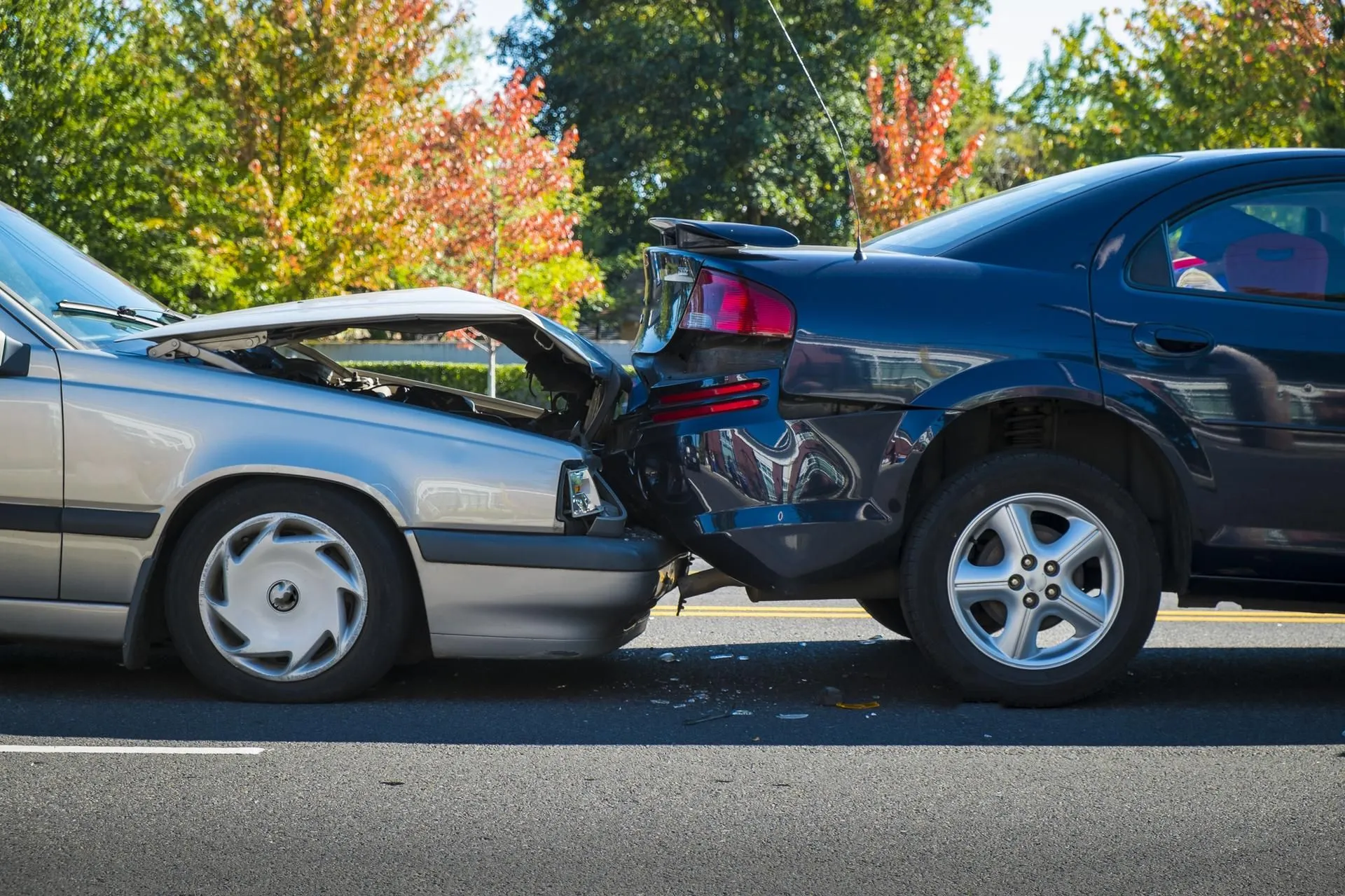 Silver car rear-ending a dark blue car on a road with severe hood damage.