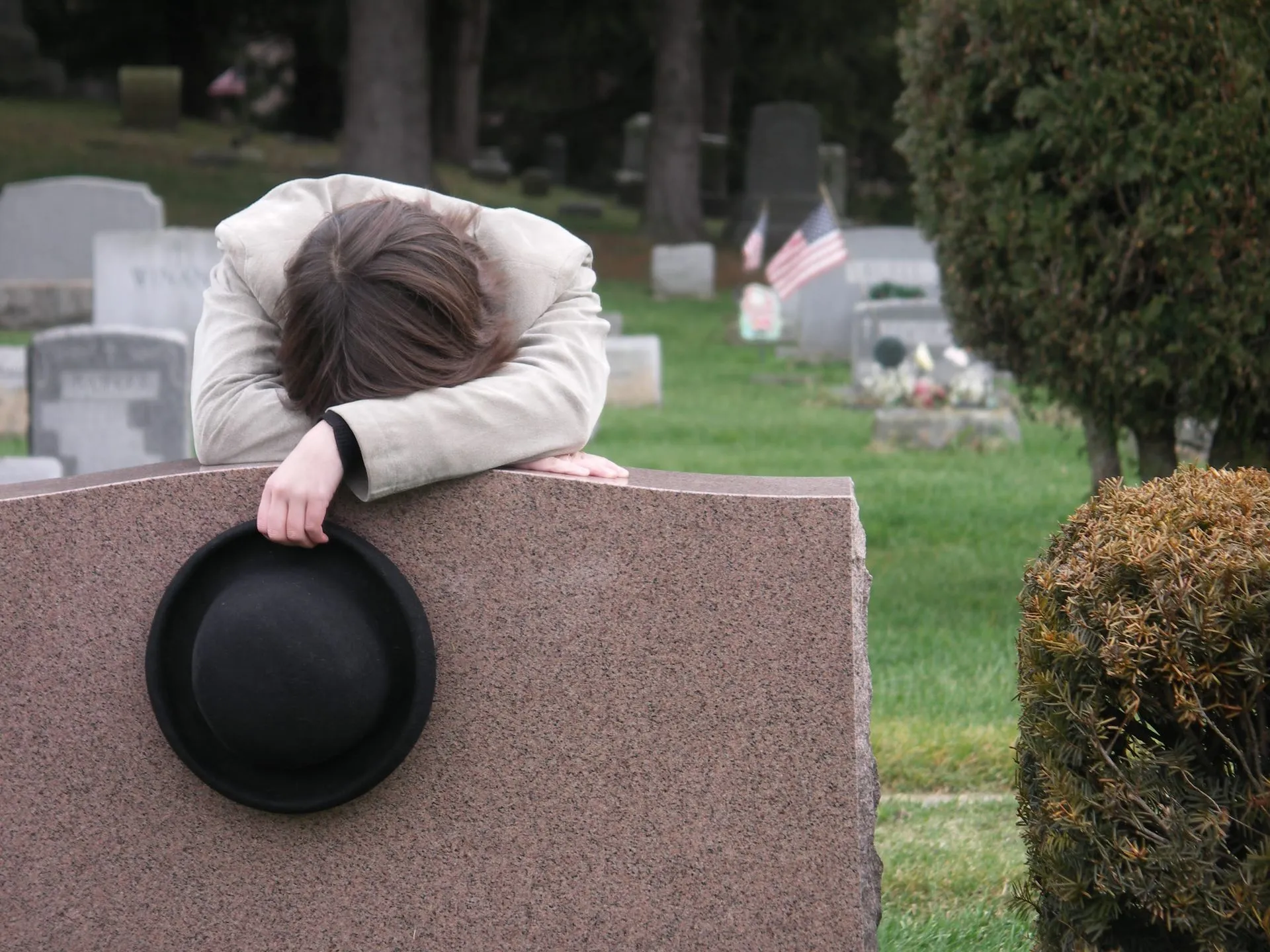 A grieving person resting on a gravestone with a hat.