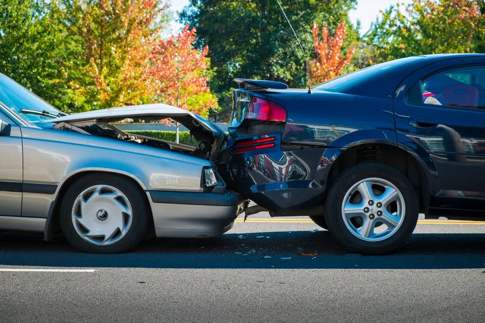 Silver car rear-ending a dark blue car on a road with severe hood damage.