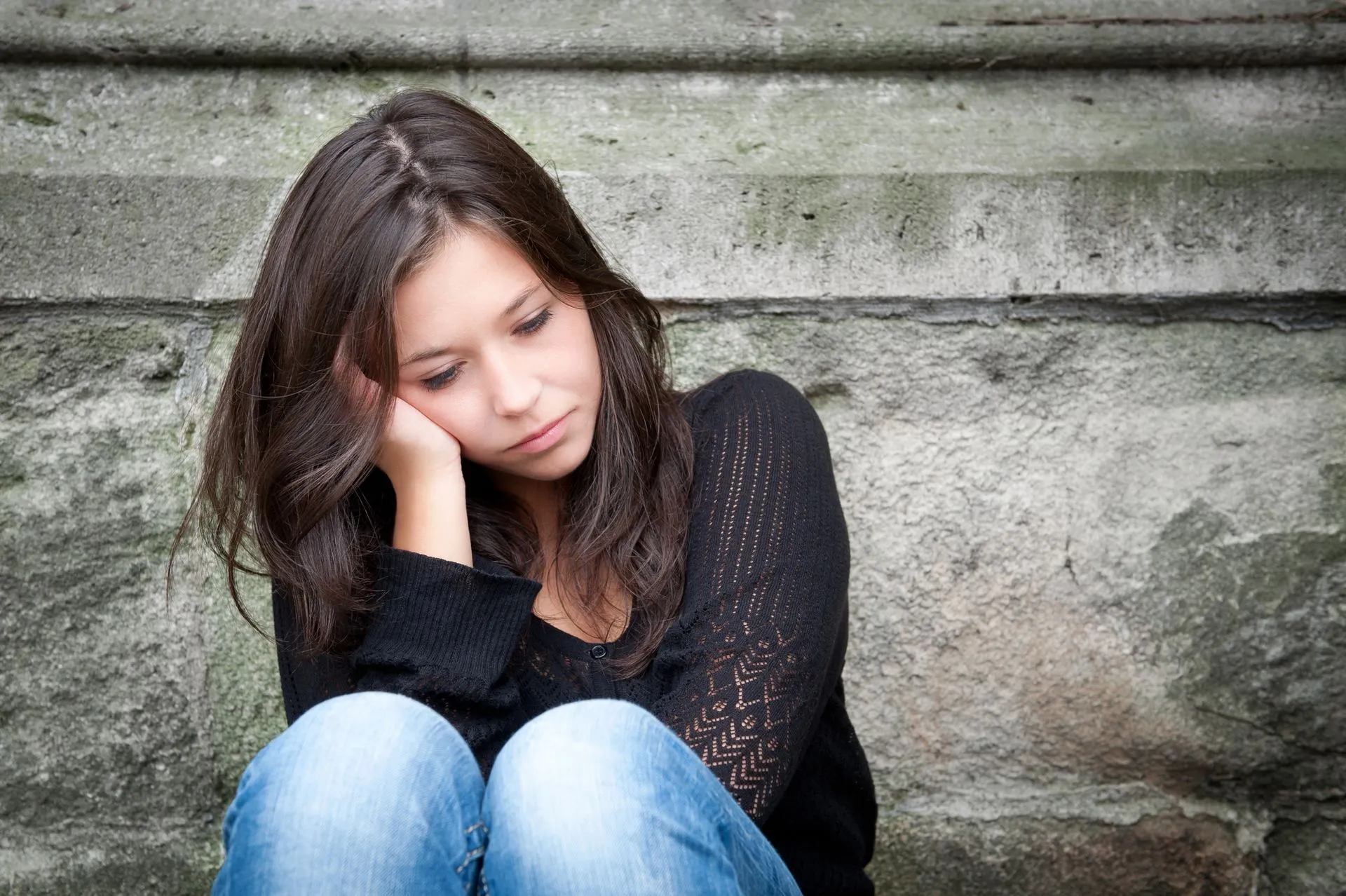 A sad young woman sitting against a stone wall.