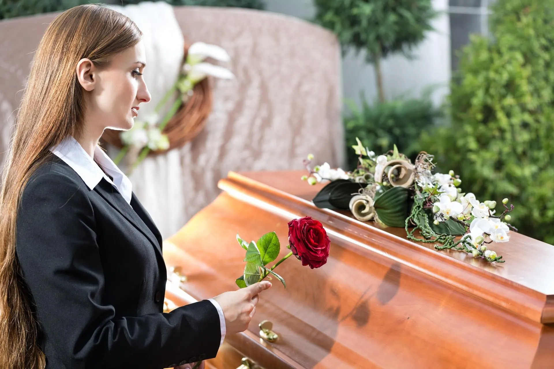 A woman holding a red rose by a wooden coffin.