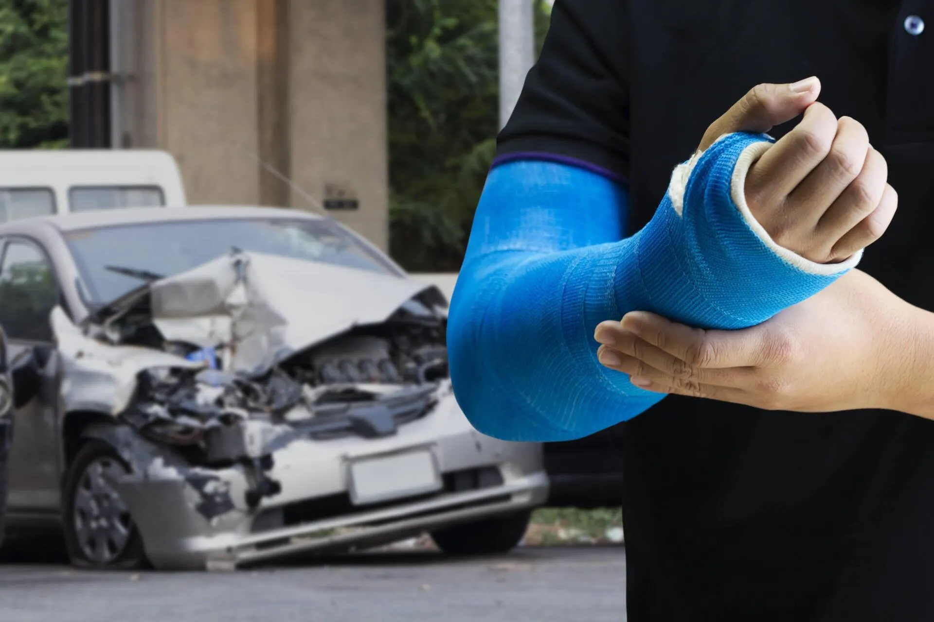 A person with a blue arm cast in front of a crashed car.