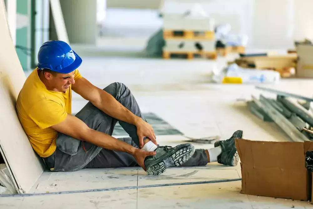 A construction worker sitting on the floor and holding his ankle.