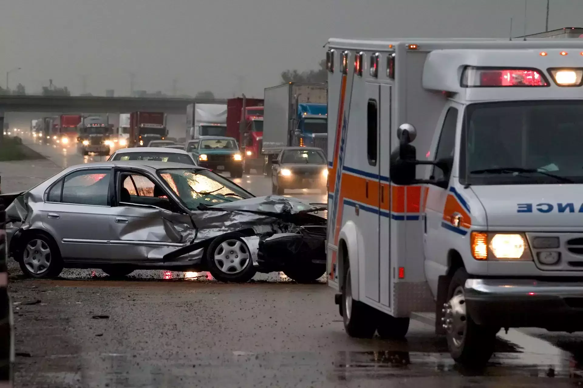 An ambulance near a heavily damaged silver car on a rainy highway.