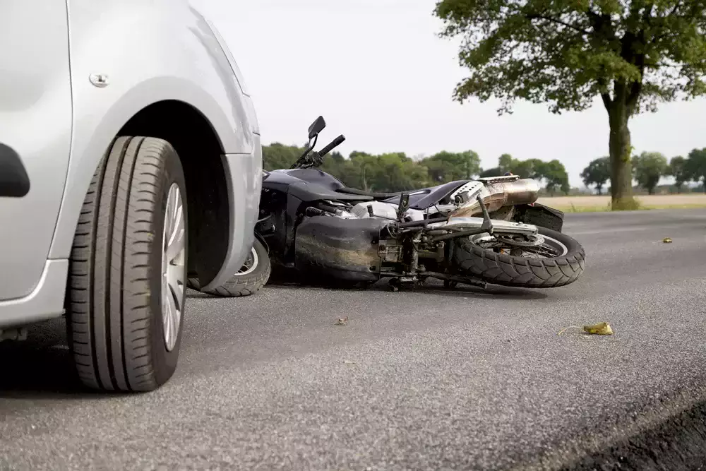Motocicleta caída en la carretera junto al neumático de un auto plateado.