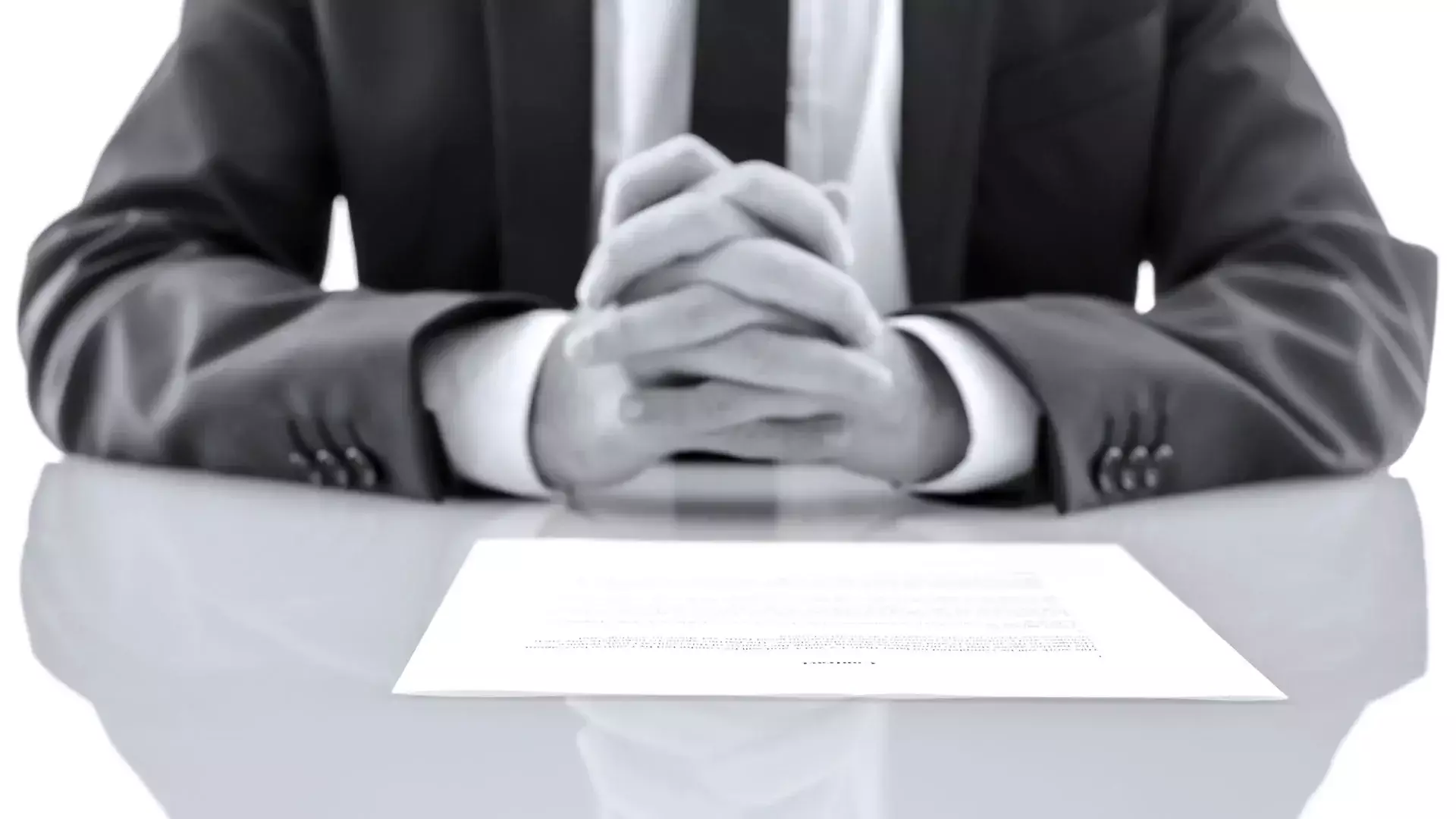 Person in a suit with clasped hands sitting at a desk behind a document.