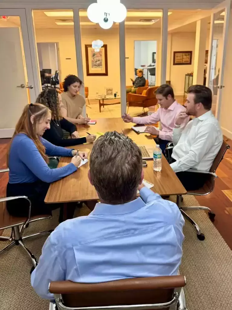 A group of six people engaged in a meeting around a wooden table in a modern office with glass walls and warm lighting.