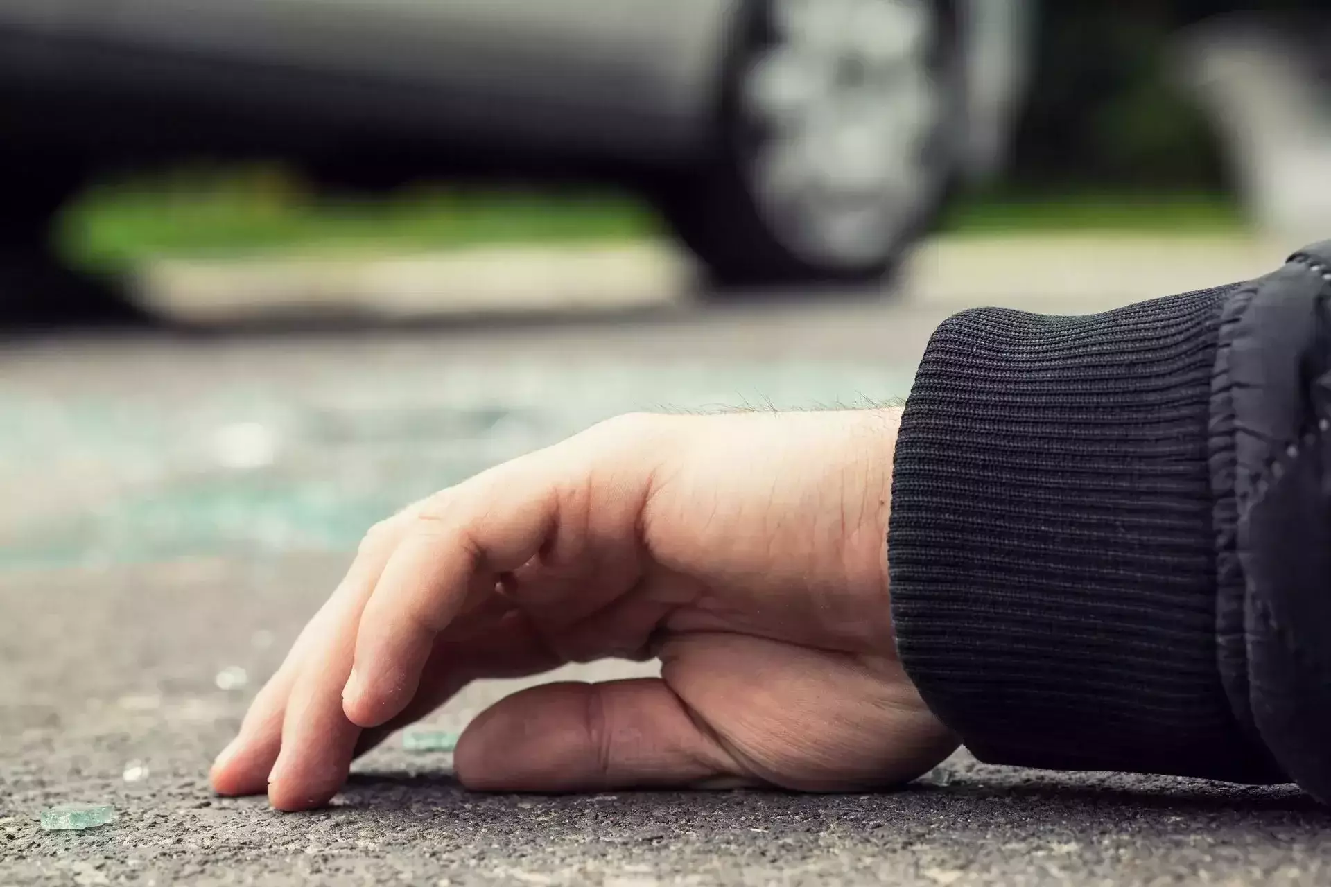 A person's hand lying on the pavement near broken glass.