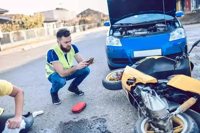 Un hombre con chaleco tomando fotos de una motocicleta accidentada y un auto azul.