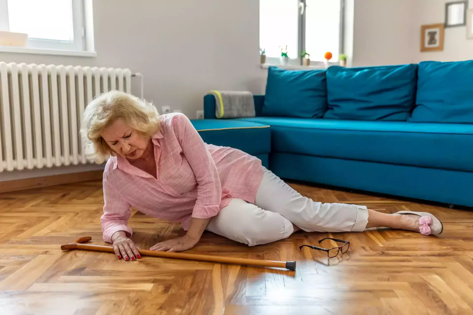 An elderly woman lying on a wooden floor near her cane and glasses.