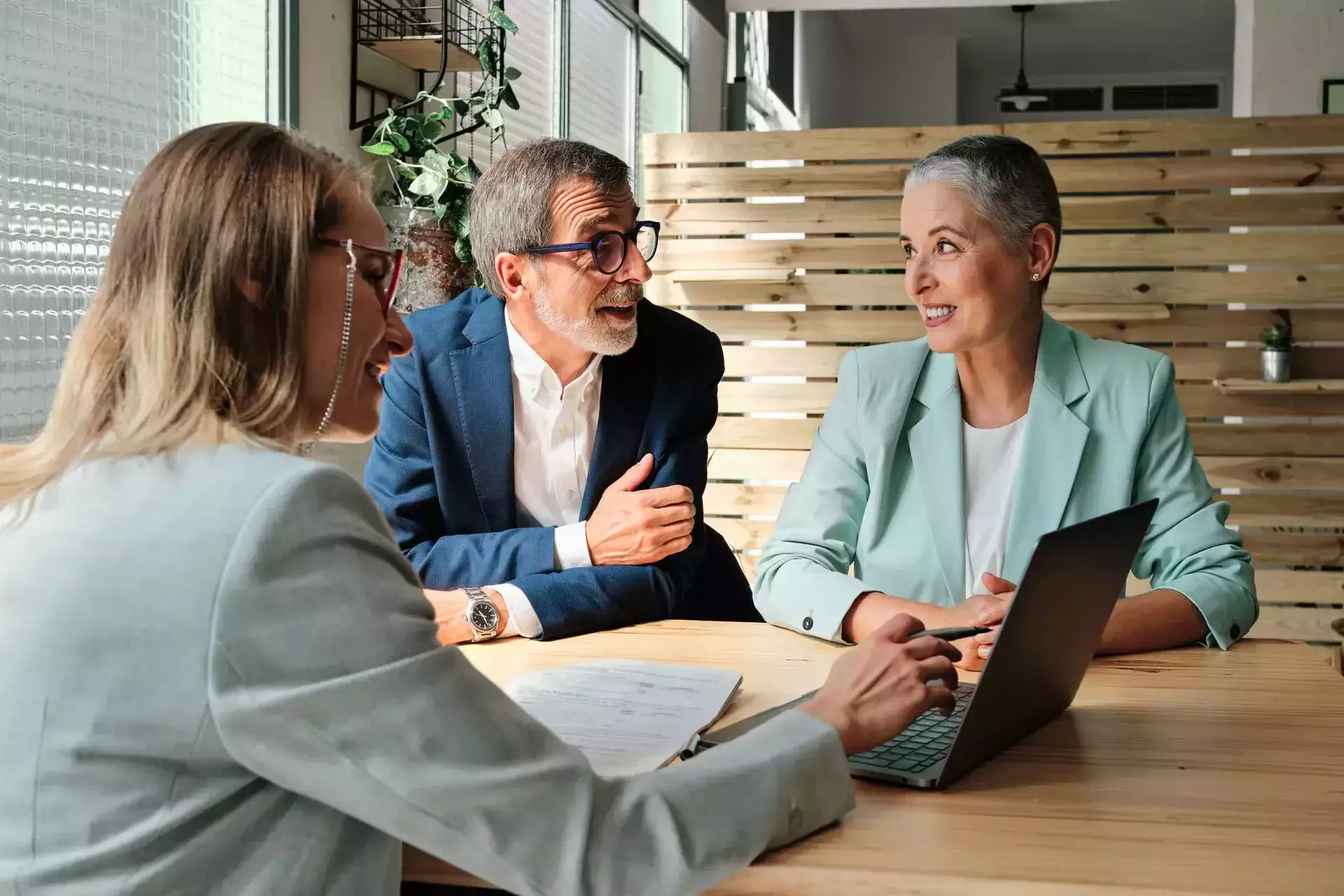 Three professionals, two women and one man, having a business discussion around a laptop at a wooden table in an office.