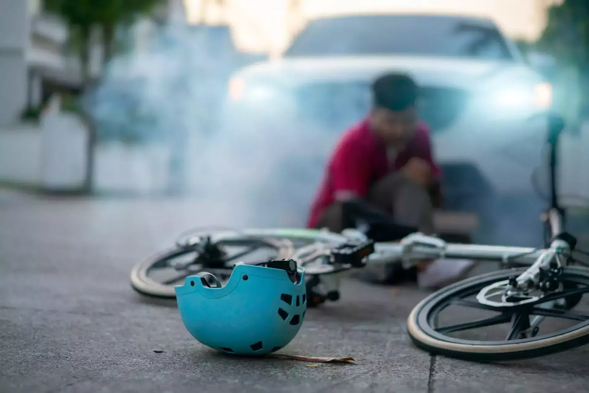 Blue helmet and fallen bicycle on road with blurred injured person and car with headlights on in the background.
