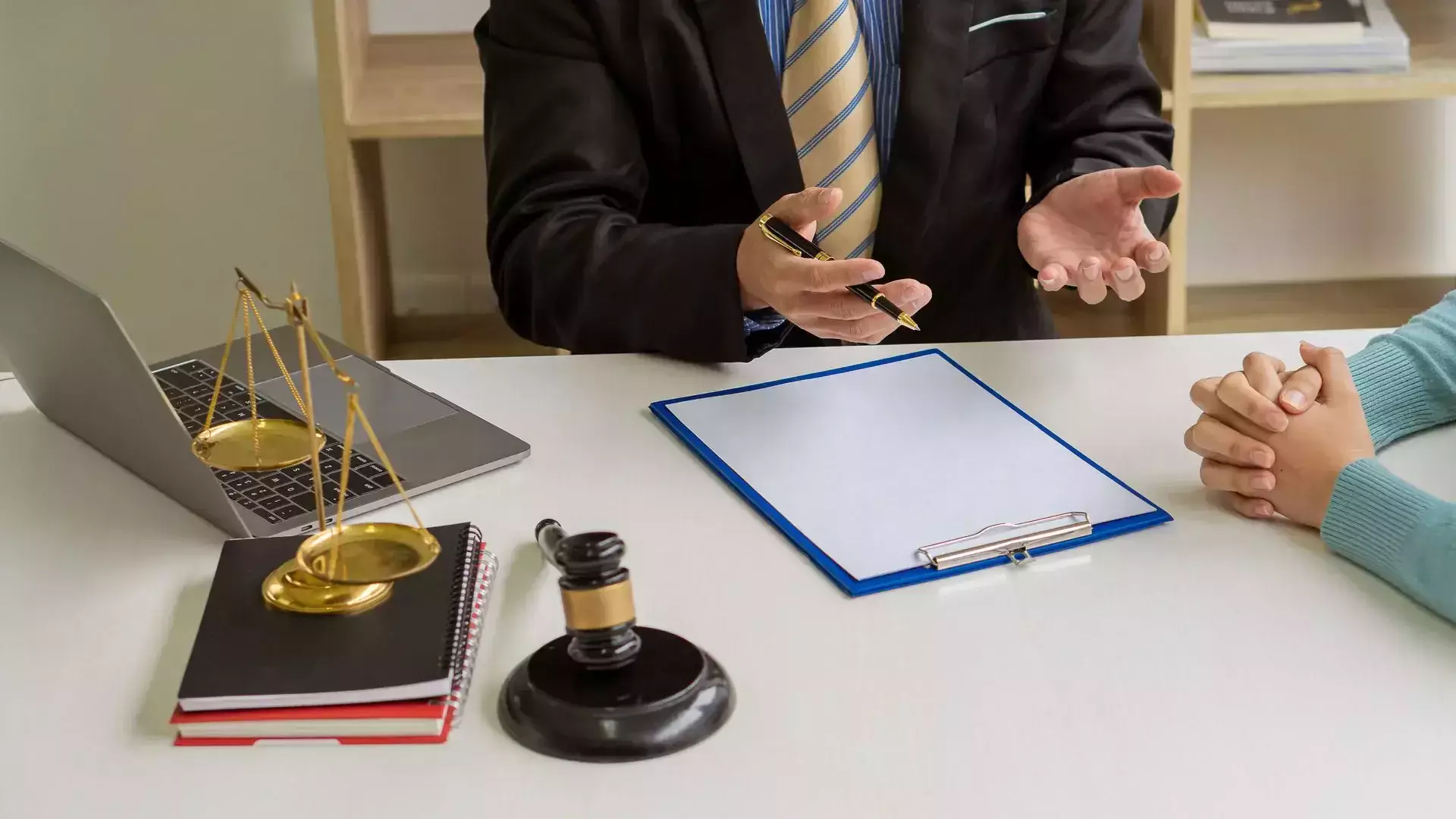 Lawyer in a suit explaining legal matters with a clipboard, laptop, scales of justice, and gavel on the desk.