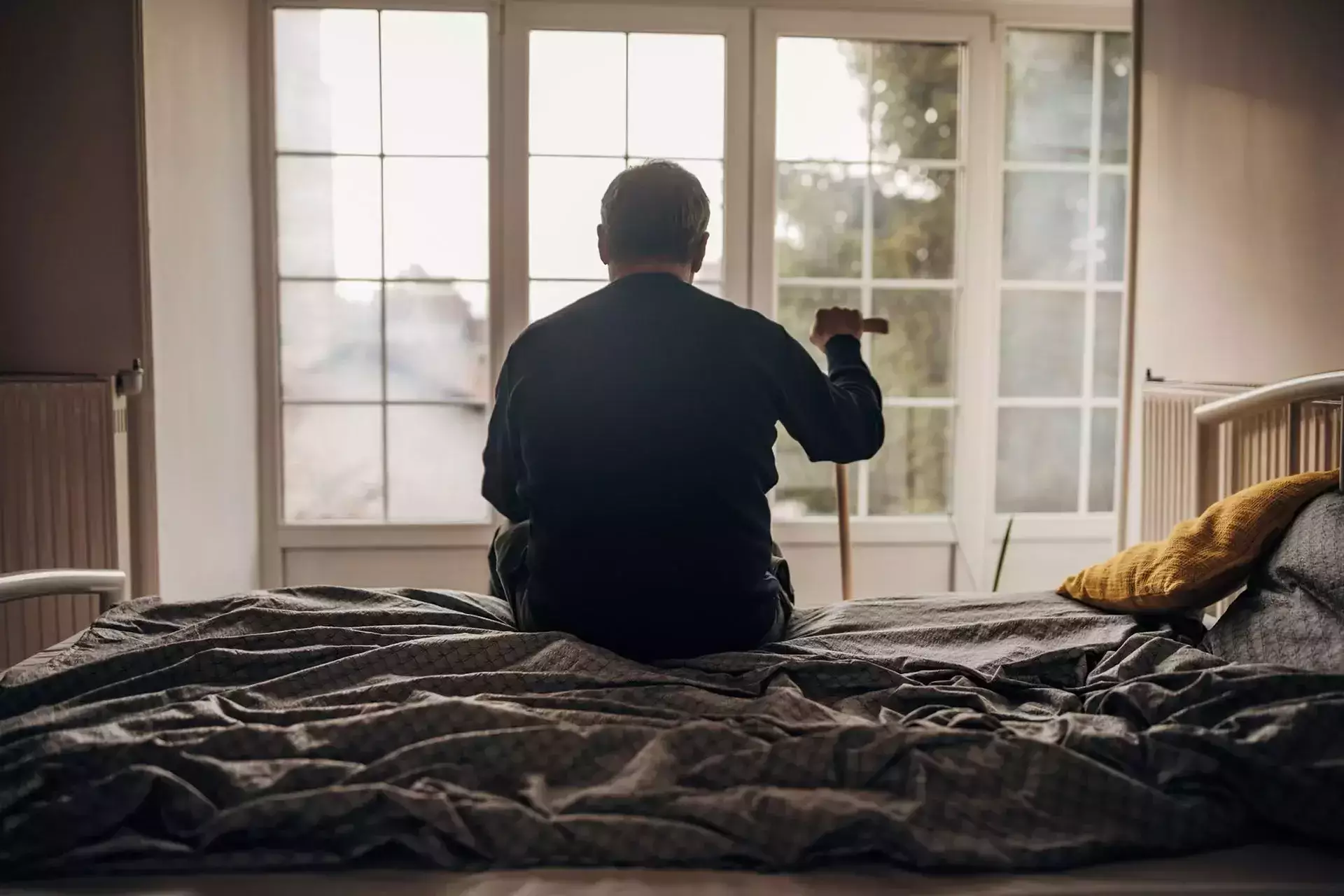 An elderly man holding a cane while sitting on the edge of a bed.
