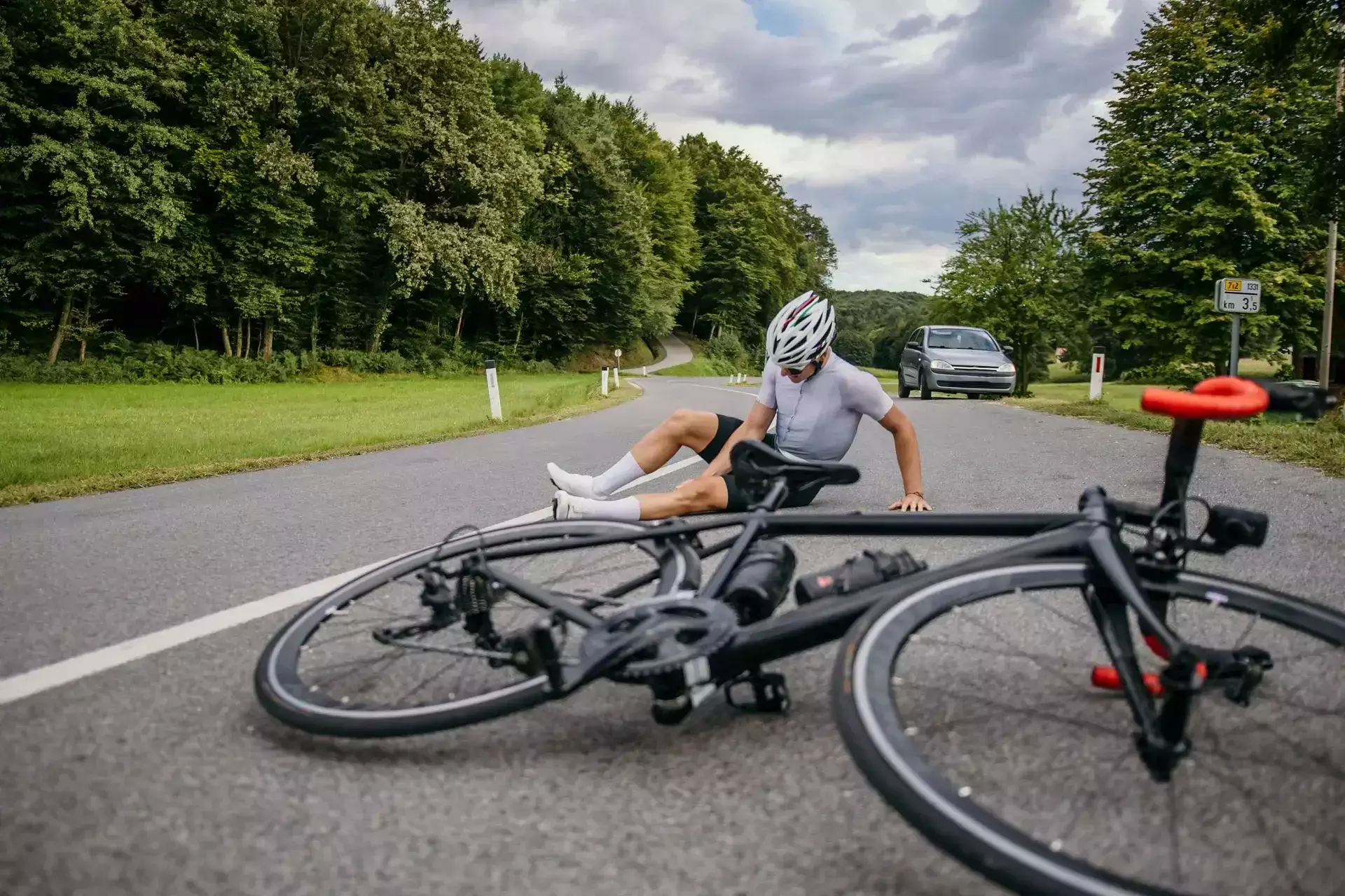 Cyclist wearing helmet sitting on road after falling beside a black road bike on a rural road with a car nearby.