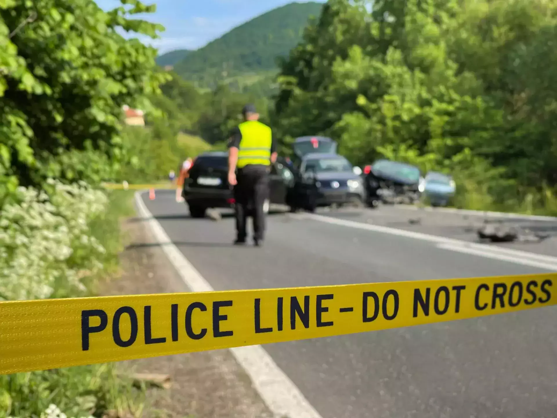 Yellow tape with 'Police Line - Do Not Cross' across a road with a car accident and officer.