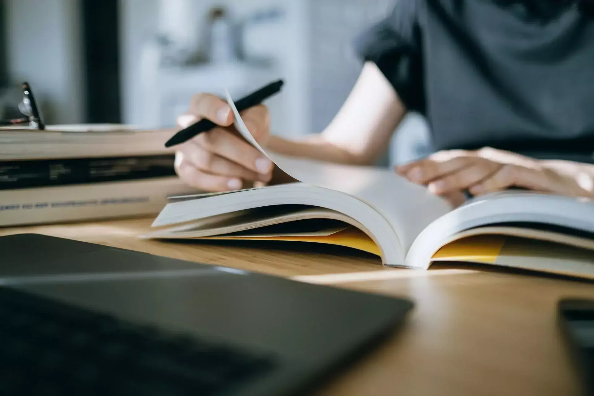 Person flipping through pages of an open book while holding a pen, with books and a laptop on a wooden table.