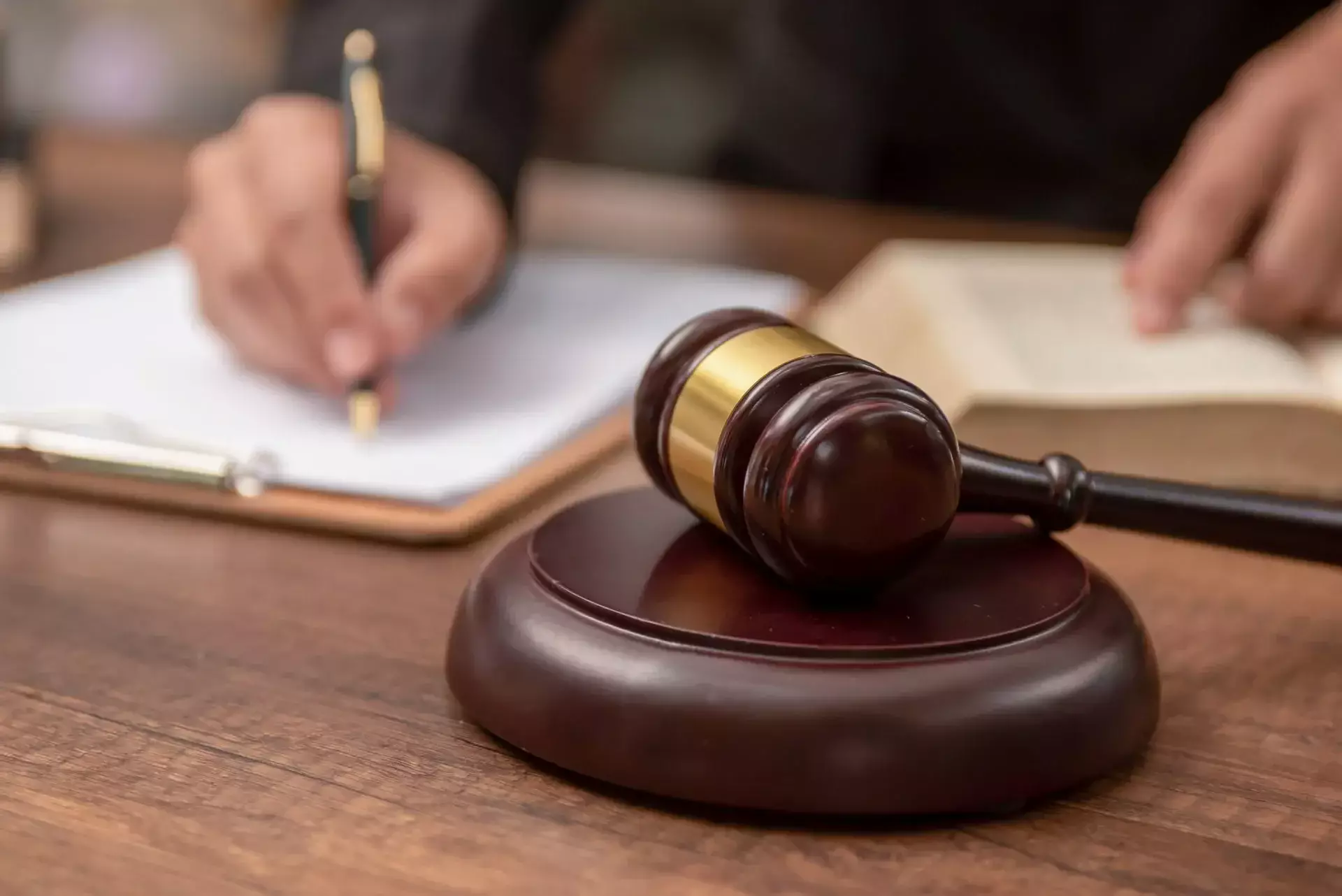 Wooden judge gavel on a desk with a person writing on a clipboard and reading a book in the background.