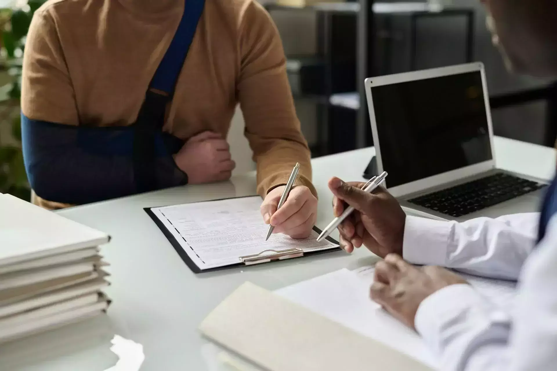 Person with arm in sling filling out a form while consulting a professional across a desk with a laptop.