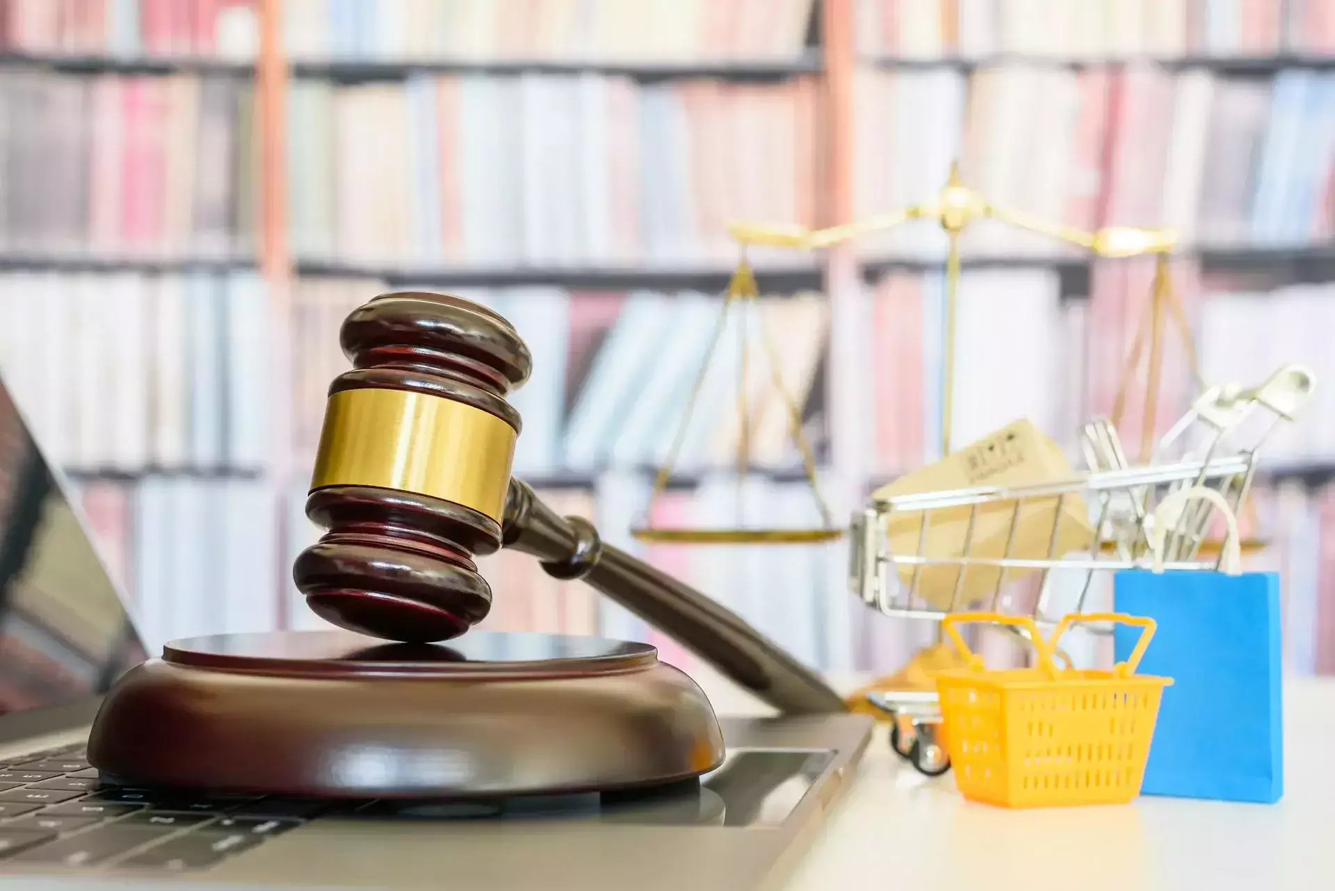 Mazo de juez sobre una computadora portátil cerca de artículos de compras en miniatura y una balanza de la justicia.