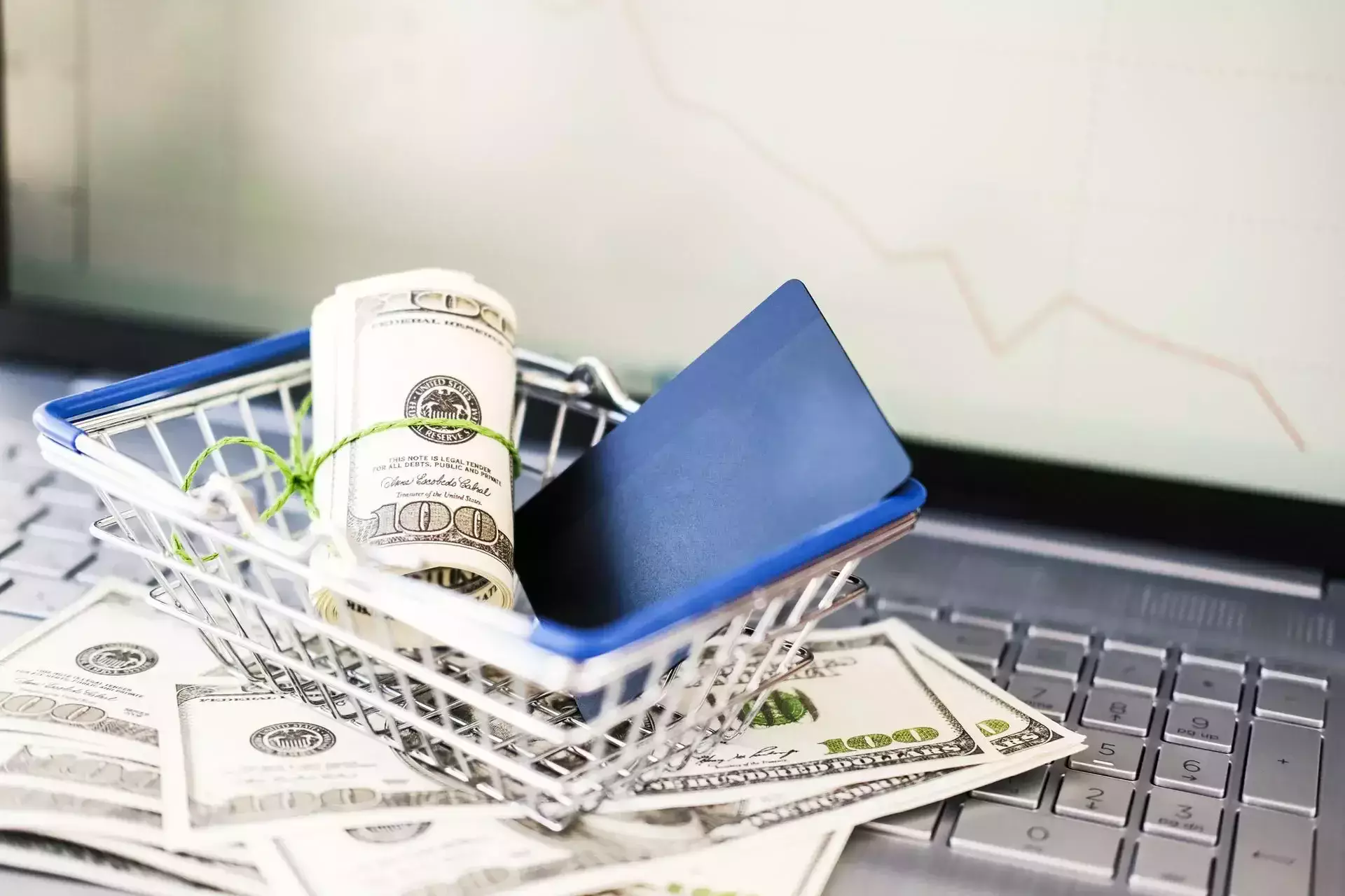 A shopping basket with cash and a card on top of a laptop.