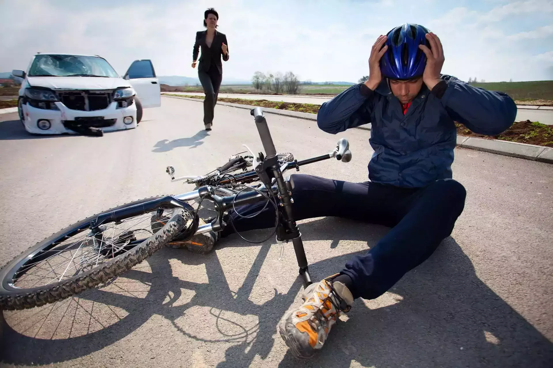 Ciclista sentado en la carretera cerca de una bicicleta destrozada y un auto dañado.