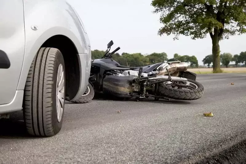 Motocicleta caída en la carretera junto al neumático de un auto plateado.
