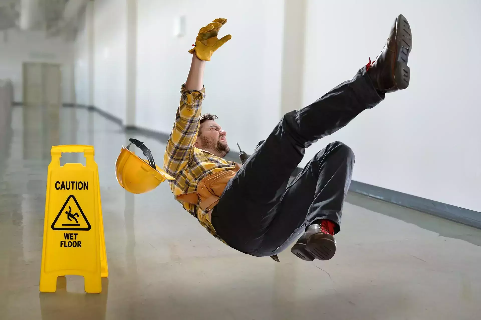 Worker slipping past a yellow sign that reads 'Caution Wet Floor' and losing his hard hat.
