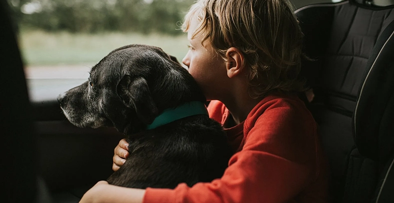 Young child sitting in a vehicle while holding a black dog.