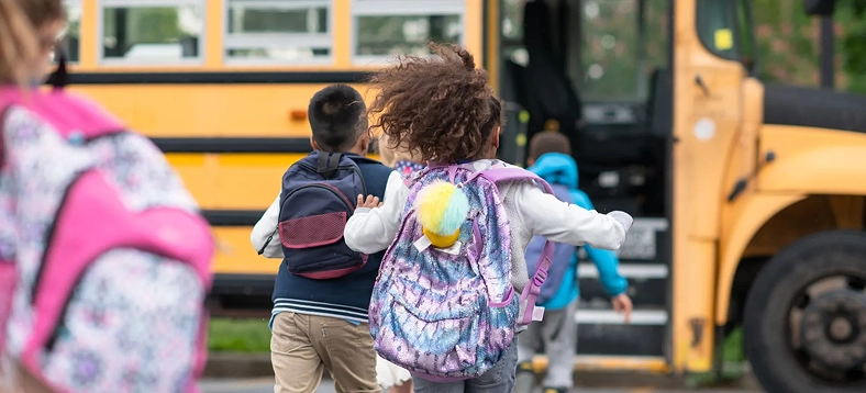 Children wearing backpacks walking toward a yellow school bus.