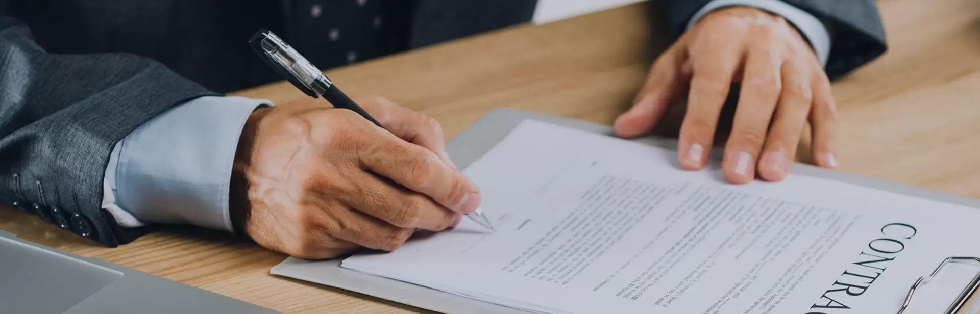 Person in suit signing a contract document on a clipboard at a wooden desk next to a laptop.