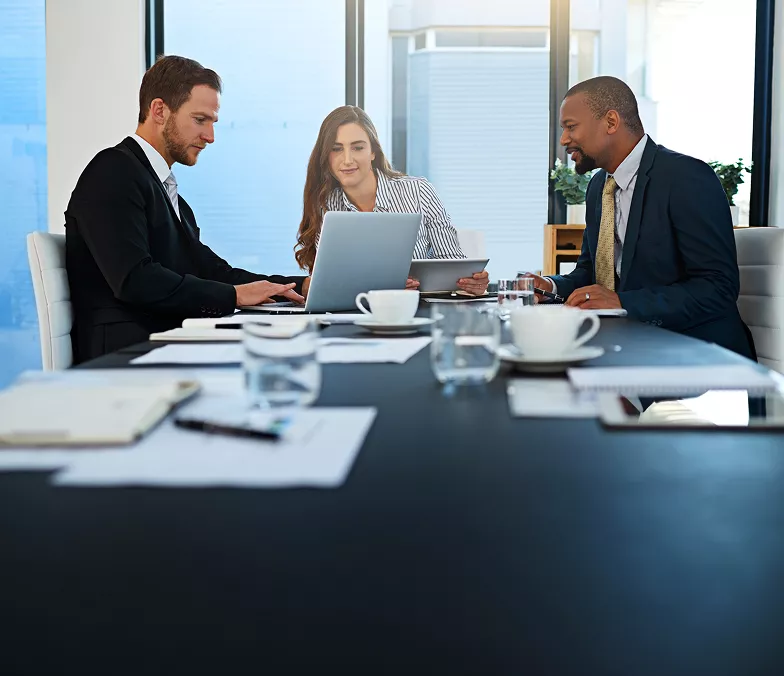 Three business professionals sitting at a conference table, working on laptops and discussing documents in a modern office setting.